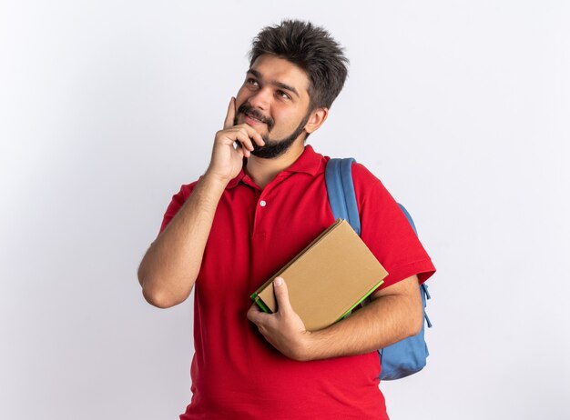 Jonge, bebaarde student in een rood poloshirt met rugzak met notitieboekjes die opkijkt met een glimlach op het gezicht, positief denkend over een witte muur