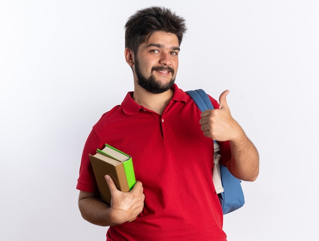 Jonge, bebaarde student in een rood poloshirt met rugzak met notitieboekjes die vrolijk lacht en duimen laat zien die over een witte muur staan