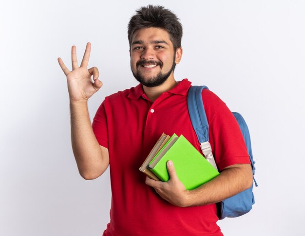 Jonge, bebaarde student in een rood poloshirt met rugzak met notitieboekjes die vrolijk lacht met een ok teken dat over een witte muur staat