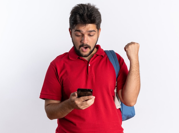 Jonge, bebaarde student in een rood poloshirt met rugzak met smartphone die er blij en verrast naar kijkt