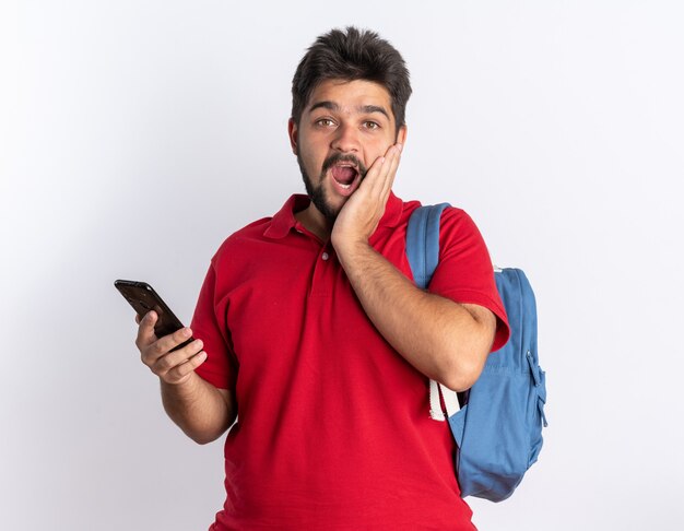 Jonge, bebaarde student in een rood poloshirt met rugzak met smartphone die er blij en verrast uitziet