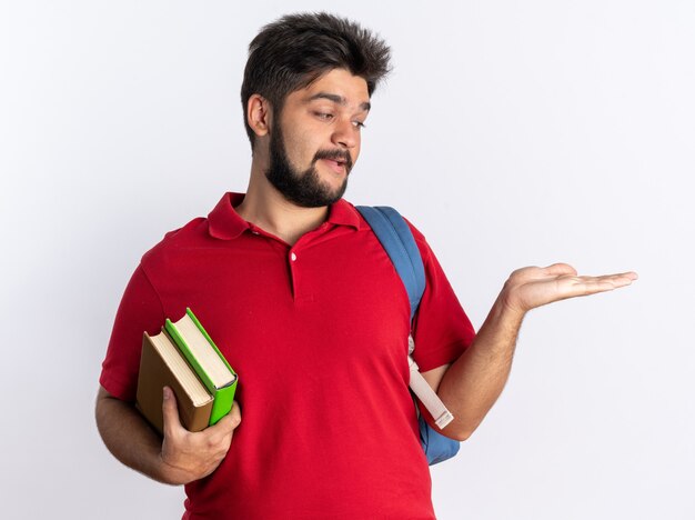 Jonge, bebaarde student man in rood poloshirt met rugzak met notebooks presenteren met arm van zijn hand kopie ruimte gelukkig en positief staande over witte muur