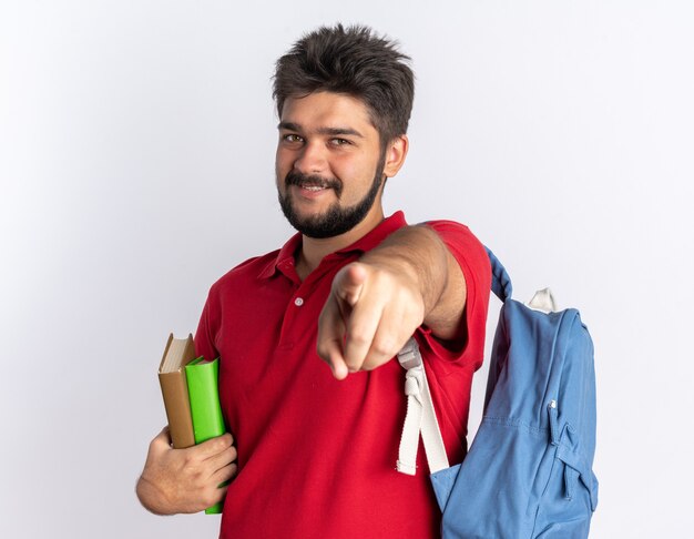 Jonge, bebaarde student man in rood poloshirt met rugzak met notebooks wijzend met wijsvinger glimlachend vrolijk gelukkig en positief staande over witte muur