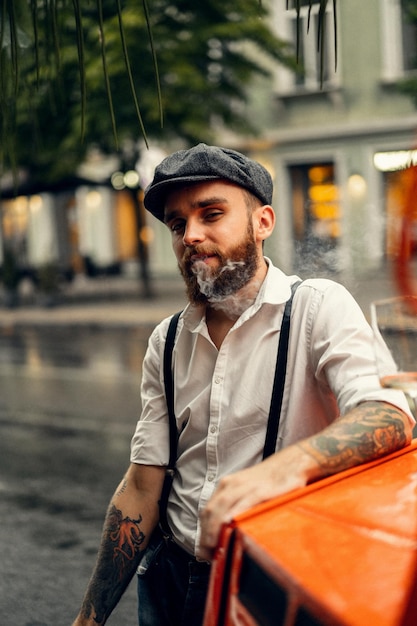 Jonge, bebaarde tattoord-man in een café op straat rookt een sigaret. Romantische man in een wit overhemd cap en bretels in de stad. Peaky Blinders. oude modieuze retro.