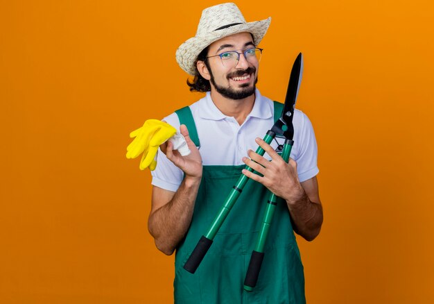 Jonge, bebaarde tuinman man met jumpsuit en hoed met heggenschaar en rubberen handschoenen kijken naar voorkant glimlachend vrolijk staande over oranje muur