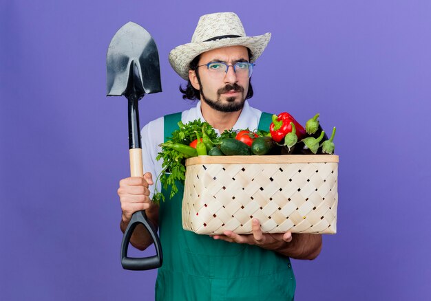 Jonge, bebaarde tuinman man met jumpsuit en hoed met krat vol groenten en schop naar voorkant kijken met ernstig gezicht staande over blauwe muur
