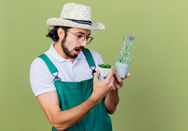 Jonge, bebaarde tuinman man met jumpsuit en hoed met potplanten kijken naar hen verrast staande over de lichtgroene muur