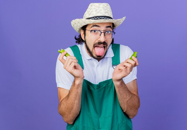 Jonge, bebaarde tuinman met jumpsuit en hoed met helften van groene hete chilipeper die uit de tong steekt, het gevoel alsof hij in zijn mond brandt en over de blauwe muur staat