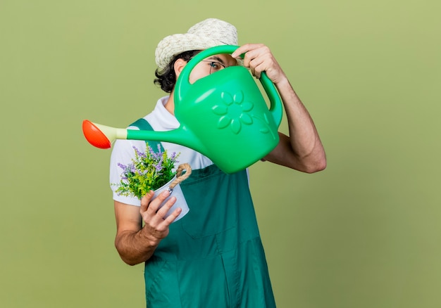 Jonge, bebaarde tuinmanmens die jumpsuit en hoed draagt die gieter en ingemaakte plant verbergt gezicht staande over lichtgroene muur