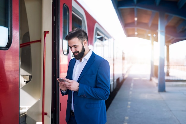 Jonge, bebaarde zakenman in elegant pak die op de metro wacht om aan het werk te gaan en zijn slimme telefoon gebruikt