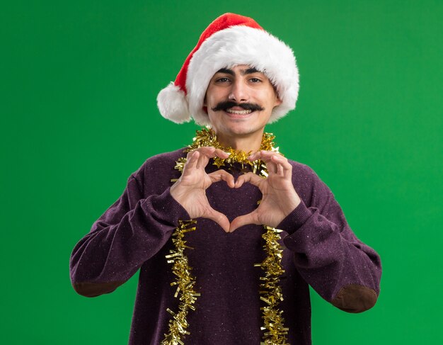 Jonge besnorde man met kerst kerstmuts met klatergoud om zijn nek kijken camera met blij gezicht lachend hart gebaar maken met vingers permanent over groene achtergrond