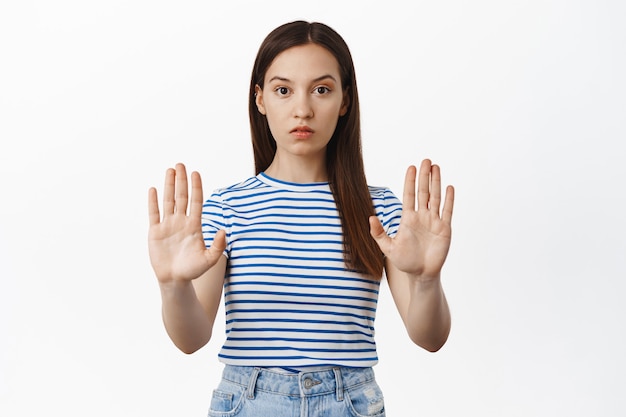 Jonge bezorgde vrouw strekt handen uit, strekt handpalmen uit in taboe, geen blokgebaar, weigert iets, keurt af en verbiedt actie, staande tegen de witte muur