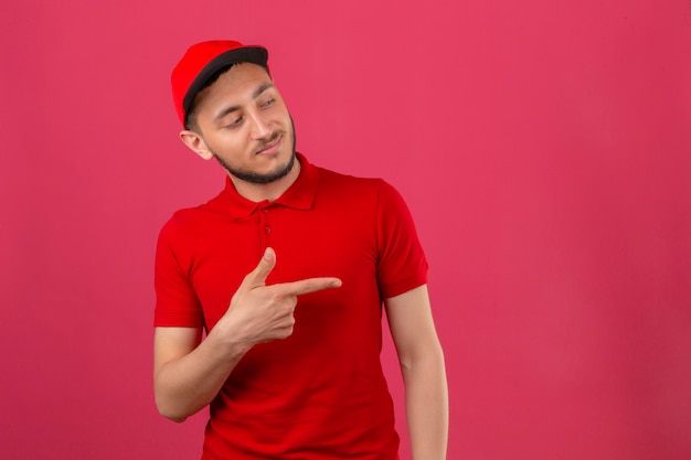 Jonge bezorger die een rood poloshirt en een pet draagt die naar de zijkant wijst om iets over geïsoleerde roze achtergrond te presenteren