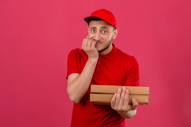 Jonge bezorger die een rood poloshirt en een pet met pizzadozen draagt die er gestrest en nerveus uitziet met de handen op de mond nagels bijten over geïsoleerde roze achtergrond