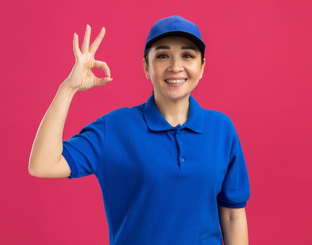 Jonge bezorger in blauw uniform en pet glimlachend vrolijk doend ok teken staande over roze muur