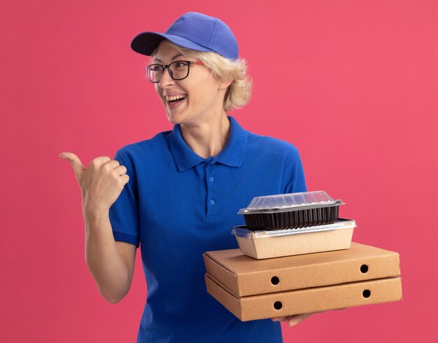 Jonge bezorger in blauw uniform en pet met bril met pizzadozen en voedselpakketten glimlachend vrolijk wijzend met wijsvinger opzij over roze muur