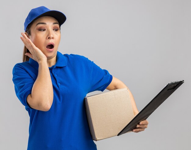 Jonge bezorger in blauw uniform en pet met kartonnen doos en klembord die er verbaasd en verrast uitziet