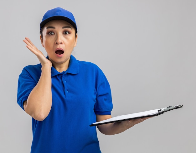 Jonge bezorger in blauw uniform en pet met klembord en pen verbaasd en verrast