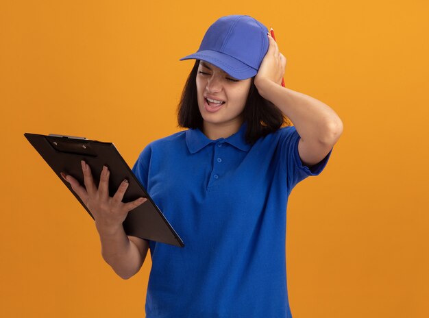 Jonge bezorger in blauw uniform en pet met klembord kijkend verward met hand op haar hoofd voor fout staande over oranje muur