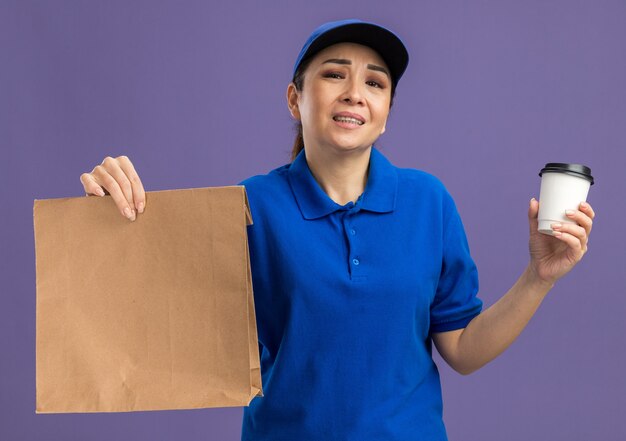 Jonge bezorger in blauw uniform en pet met papieren pakje en papieren beker geërgerd met een droevige uitdrukking die over de paarse muur staat