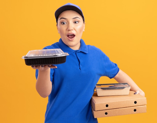 Jonge bezorger in blauw uniform en pet met pizzadozen en voedselpakketten kijken naar dozen verbaasd en verrast staande over oranje muur