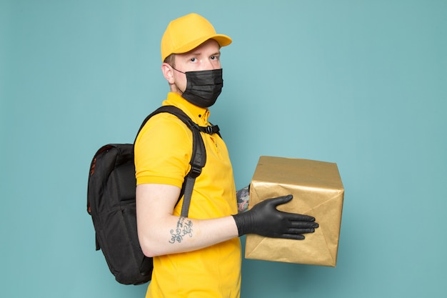 jonge bezorger in gele polo gele pet witte jeans rugzak en zwart steriel masker met een doos op blauw