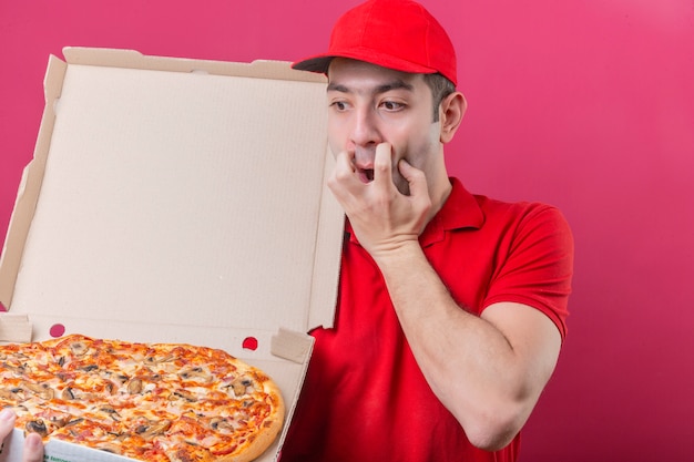 Jonge bezorger in rood poloshirt en pet permanent met doos verse pizza kijken ernaar geschokt bang over geïsoleerde roze achtergrond