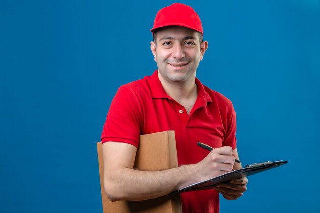 Jonge bezorger in rood poloshirt en pet permanent met klembord en pen houden pizzadoos camera kijken met glimlach op gezicht over geïsoleerde blauwe achtergrond