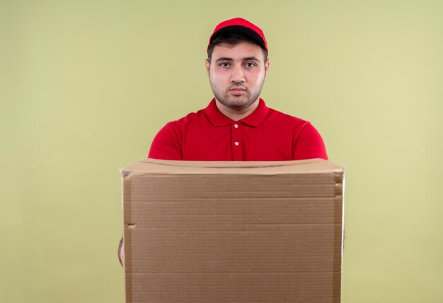 Jonge bezorger in rood uniform en GLB met doos pakket met ernstig gezicht staande over groene muur