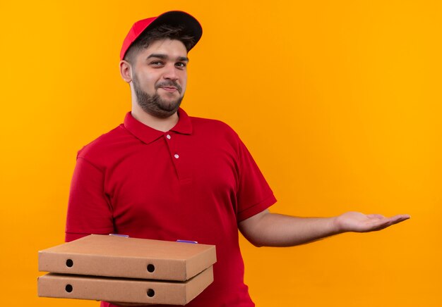 Jonge bezorger in rood uniform en pet bedrijf stapel pizzadozen presenteren met arm oh zijn hand kopie ruimte