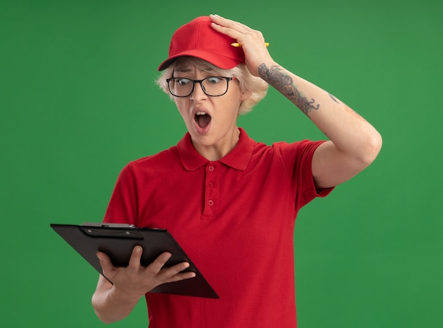 Jonge bezorger in rood uniform en pet die een bril met klembord draagt en ernaar kijkt, verward met de hand op haar hoofd voor een fout die over de groene muur staat