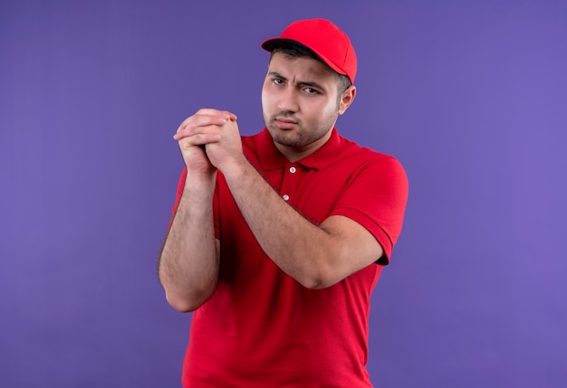 Jonge bezorger in rood uniform en pet die groepswerkgebaar hand in hand samen met ernstig gezicht maken dat zich over purpere muur bevindt