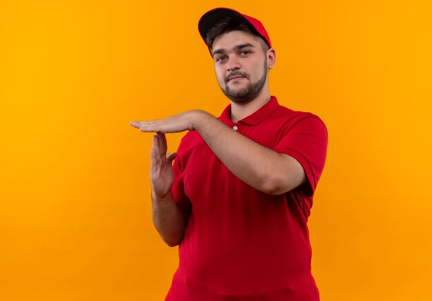 Jonge bezorger in rood uniform en pet maken time-out gebaar met handen camera kijken