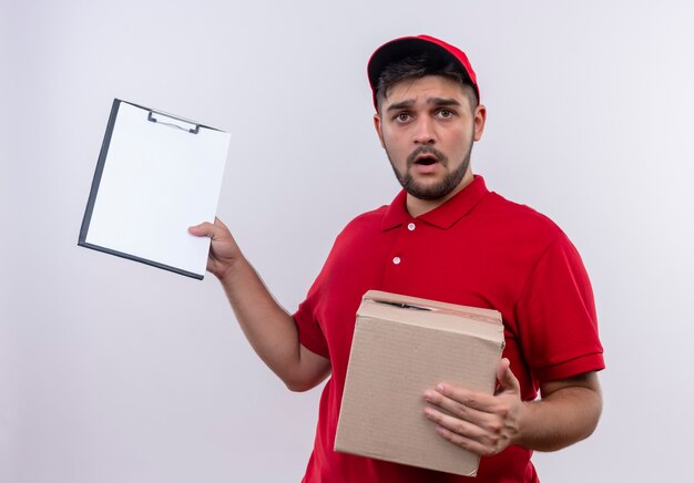 Jonge bezorger in rood uniform en pet met doos pakket met klembord met blanco pagina's die er verward en erg angstig uitzien