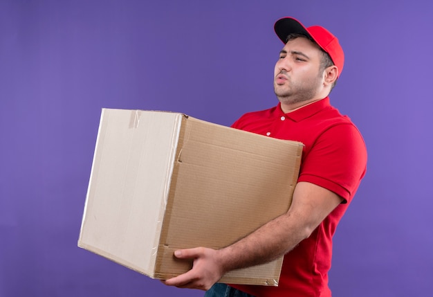 Jonge bezorger in rood uniform en pet met een grote kartonnen doos die er ziek uitziet en lijdt aan zwaar gewicht dat over de paarse muur staat