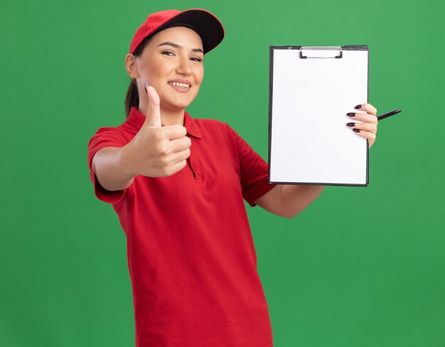 Jonge bezorger in rood uniform en pet met klembord met blanco pagina's kijken naar voorkant glimlachend vrolijk tonen duimen omhoog staande over groene muur