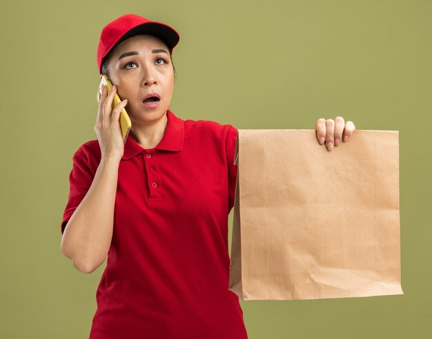 Jonge bezorger in rood uniform en pet met papieren pakket die verward en verrast kijkt terwijl ze op een mobiele telefoon praat die over een groene muur staat