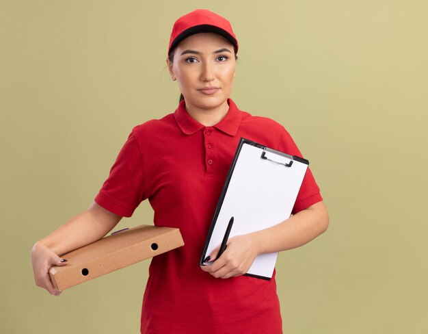 Jonge bezorger in rood uniform en pet met pizzadoos en klembord met balnk pagina's kijken naar voorkant met ernstig gezicht staande over groene muur