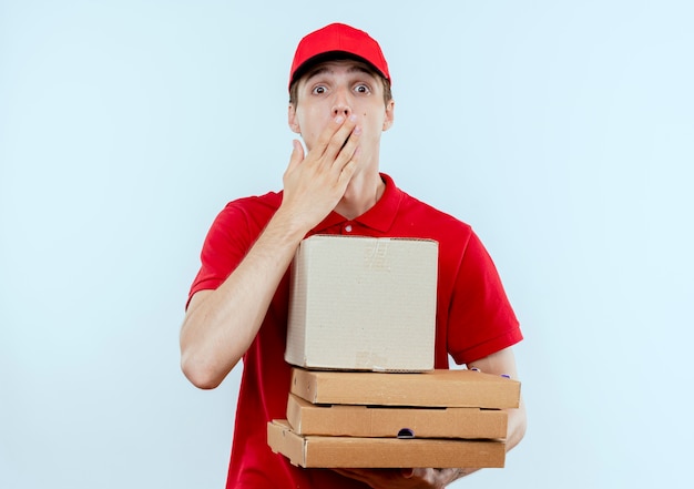 Jonge bezorger in rood uniform en pet met pizzadozen en doospakket op zoek verrast en verbaasd staande over witte muur