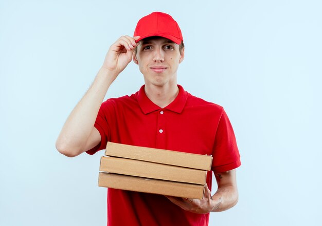 Jonge bezorger in rood uniform en pet met pizzadozen op zoek naar de voorkant met zelfverzekerde uitdrukking staande over witte muur