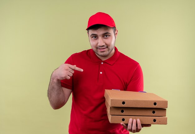 Jonge bezorger in rood uniform en pet met pizzadozen wijzend met de vinger naar hen glimlachend zelfverzekerd staande over groene muur