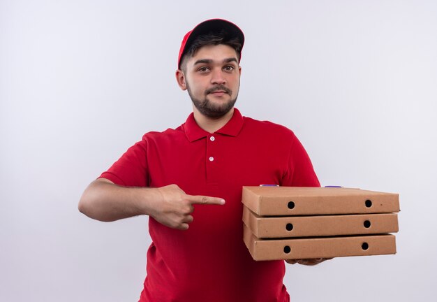 Jonge bezorger in rood uniform en pet met pizzadozen wijzend met wijsvinger vriendelijk glimlachen