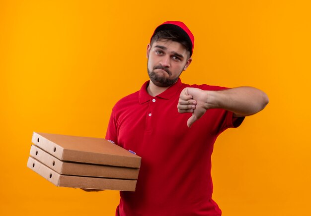 Jonge bezorger in rood uniform en pet met stapel pizzadozen met duimen naar beneden met droevige uitdrukking op gezicht