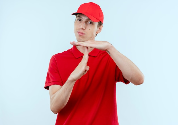 Jonge bezorger in rood uniform en pet op zoek naar de voorkant met ernstig gezicht waardoor time-out gebaar met handen over witte muur