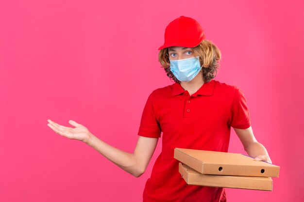 Jonge bezorger in rood uniform met medisch masker met pizzadozen glimlachend vrolijk presenteren en wijzen met handpalm kijkend naar de camera over geïsoleerde roze achtergrond