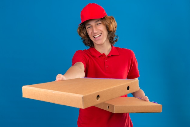 Gratis foto jonge bezorger in rood uniform pizzadozen uitrekken glimlachend vriendelijk staande over geïsoleerde blauwe achtergrond