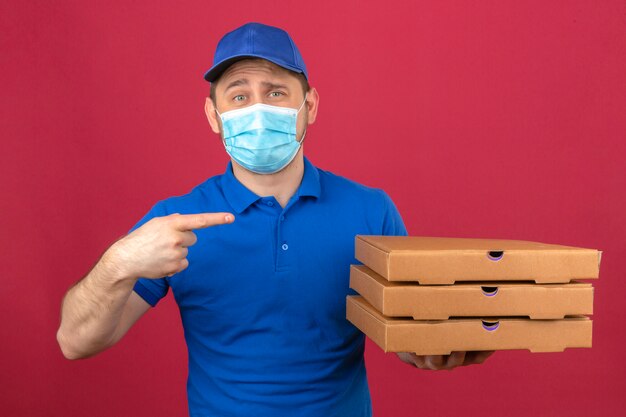 Jonge bezorger met blauw poloshirt en pet in medisch masker met stapel pizzadozen wijzende vinger naar hen kijken camera met glimlach staande over geïsoleerde roze achtergrond