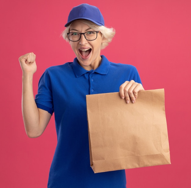 Jonge bezorgvrouw in blauw uniform en pet die een bril draagt die een papieren pakket draagt, vrolijk gebalde vuist blij en opgewonden over roze muur lacht