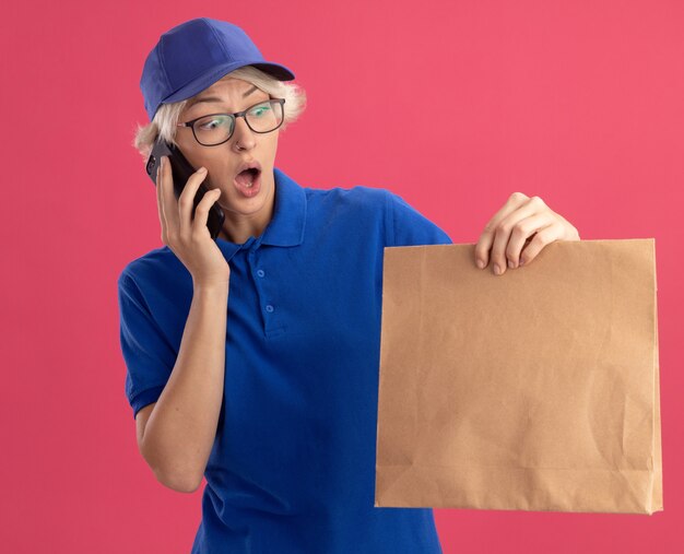 Jonge bezorgvrouw in blauw uniform en pet met papieren pakket op zoek verrast en verbaasd tijdens het praten over de mobiele telefoon over de roze muur