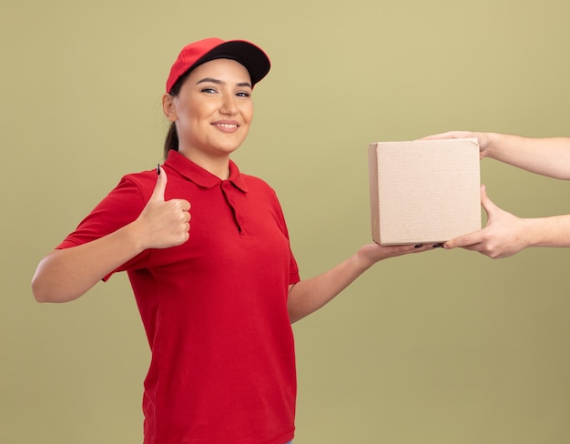 Jonge bezorgvrouw in rood uniform en pet die kartonnen doos geeft aan een klant die vriendelijk lacht toont duimen omhoog die zich over groene muur bevinden